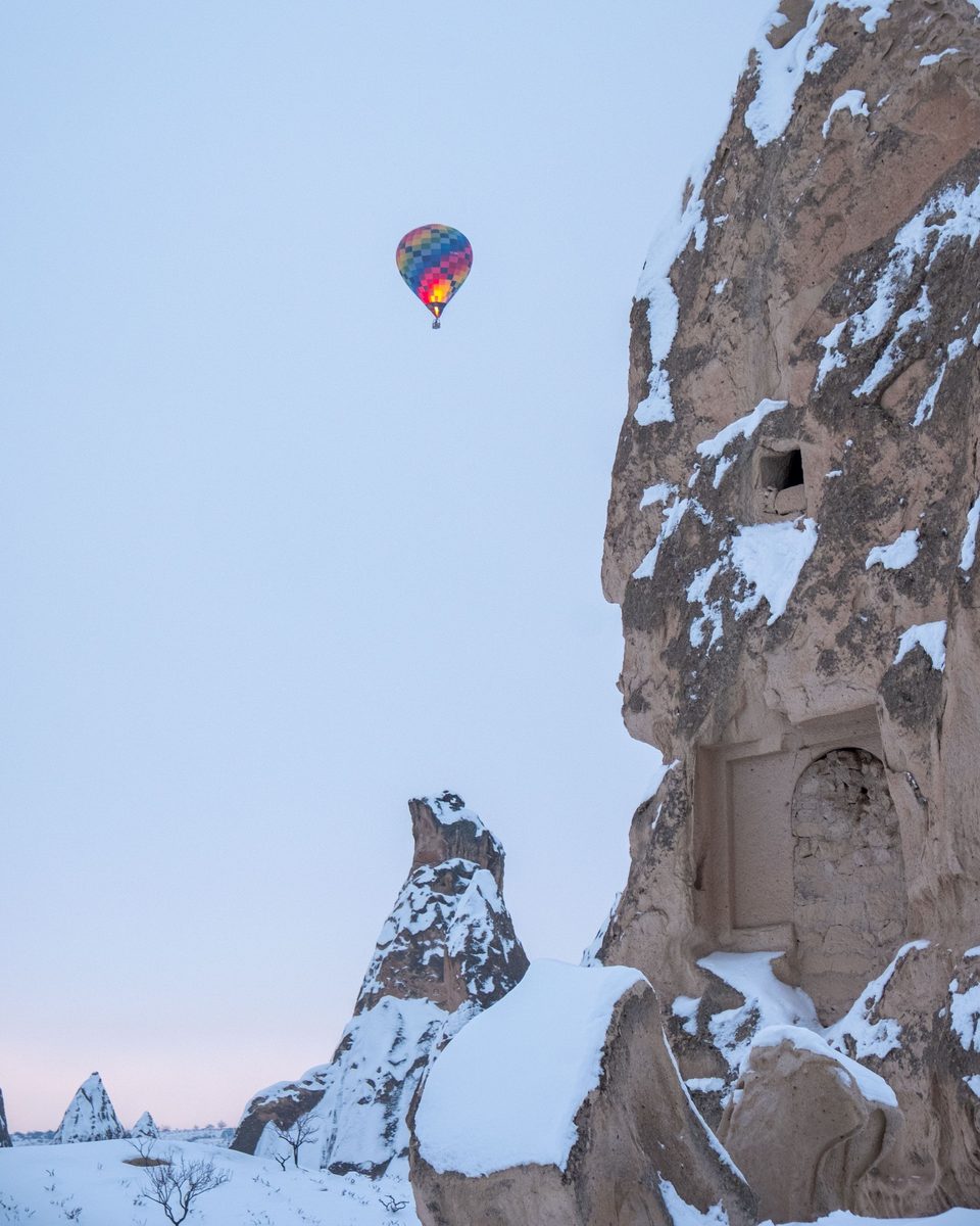 A single hot-air balloon rising at dawn beside a snow-covered rock with a carved doorway and window in Cappadocia