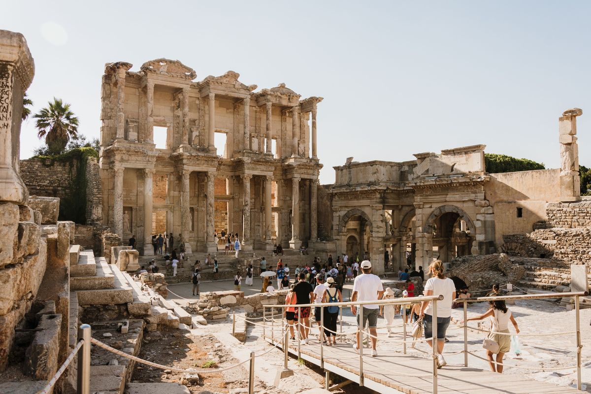 Visitors crossing a wooden boardwalk toward the Library of Celsus and the Gate of Mazeus and Mithridates on a summer day at Ephesus