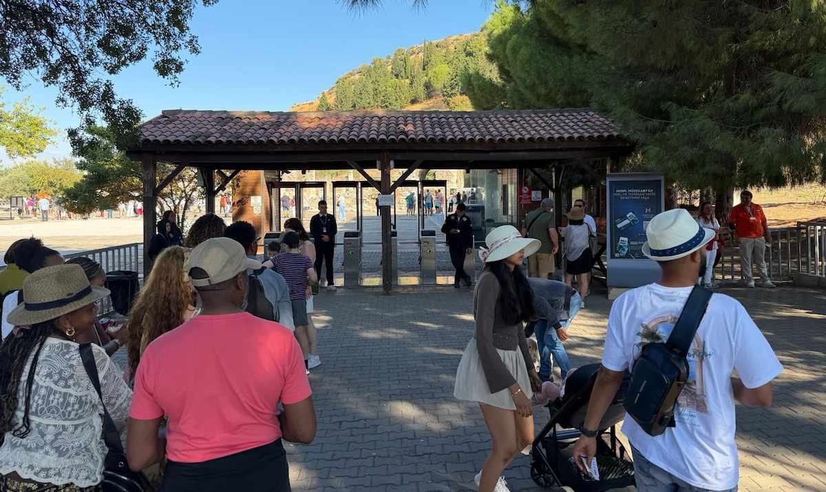 Visitors walking toward the turnstiles under the tiled roof of the Upper Gate entrance at Ephesus, with pines and the hillside behind
