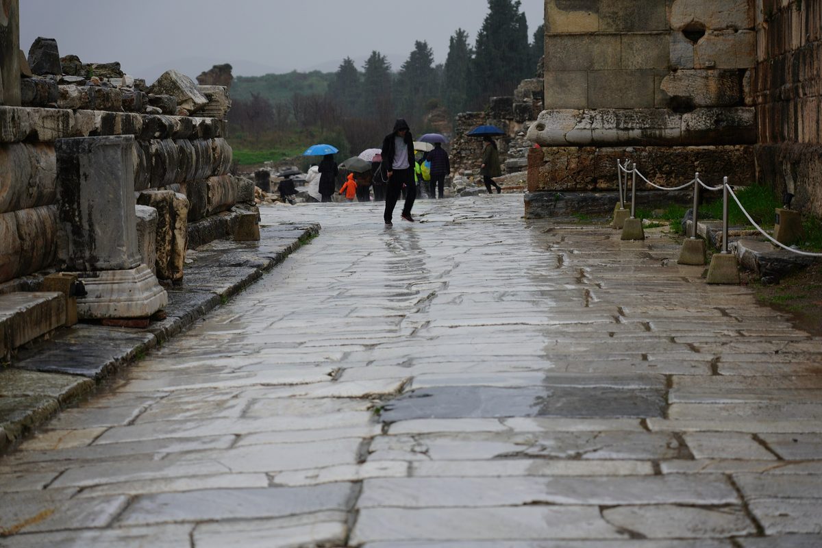 The wet marble paving of Marble Street at Ephesus on a rainy winter day, with a few visitors under umbrellas and cypress trees in the mist