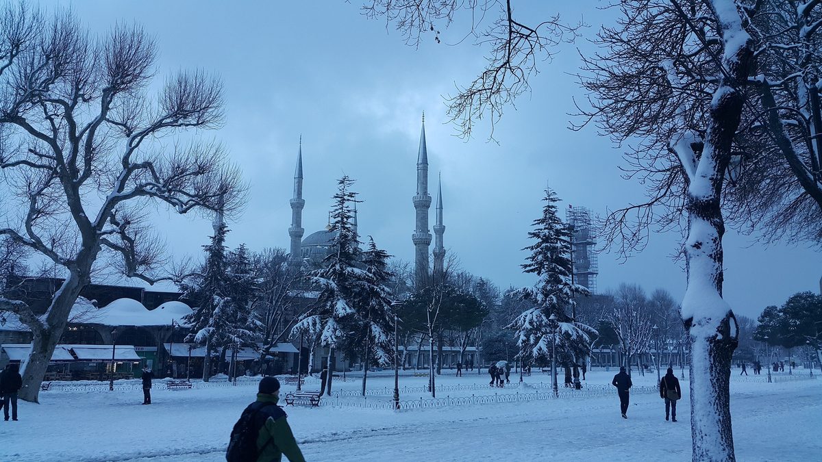 The Blue Mosque and its minarets seen across snow-covered Sultanahmet Park in Istanbul, with bare plane trees and a few people walking