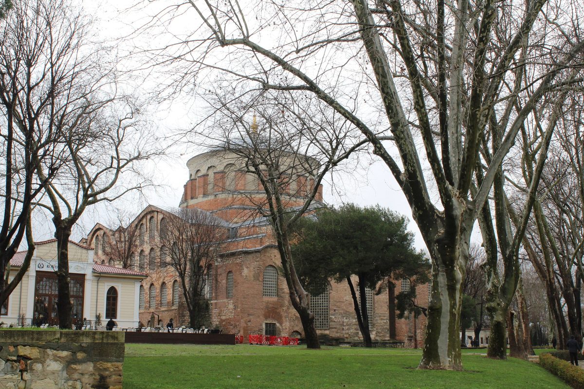 The red-brick dome of Hagia Irene in the first courtyard of Topkapı Palace on a gray winter day, framed by bare plane trees over green lawns
