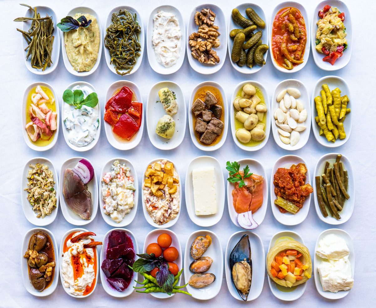 Rows of small white plates of Turkish meze, including stuffed mussels, roasted peppers, yogurt dips and pickles, on a white tablecloth