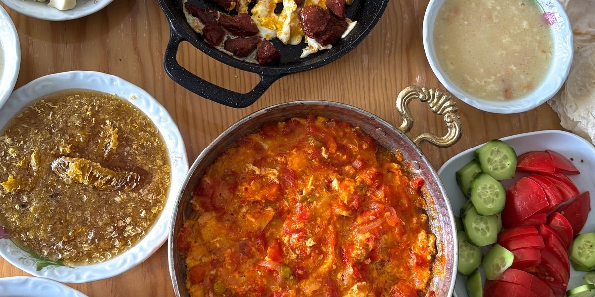 A Turkish breakfast table seen from above: menemen in a copper pan, honey on the comb, sucuk with fried eggs, tomatoes and cucumbers